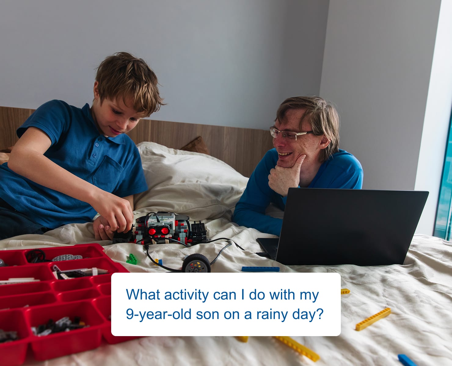 Father and son on a bed building a robot together, with a laptop open showing a Perplexity-style chat saying: What activity can I do with my 9-year-old son on a rainy day?